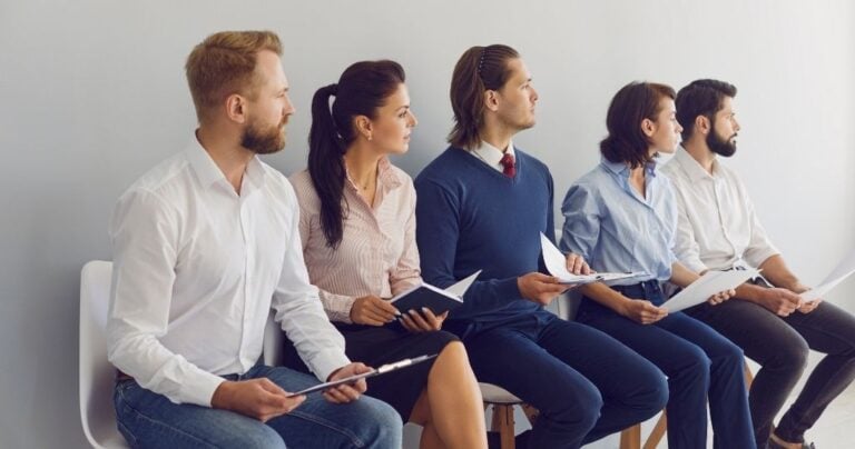 A group of candidates sitting in a row on chairs, holding resumes and notebooks while waiting for an interview.