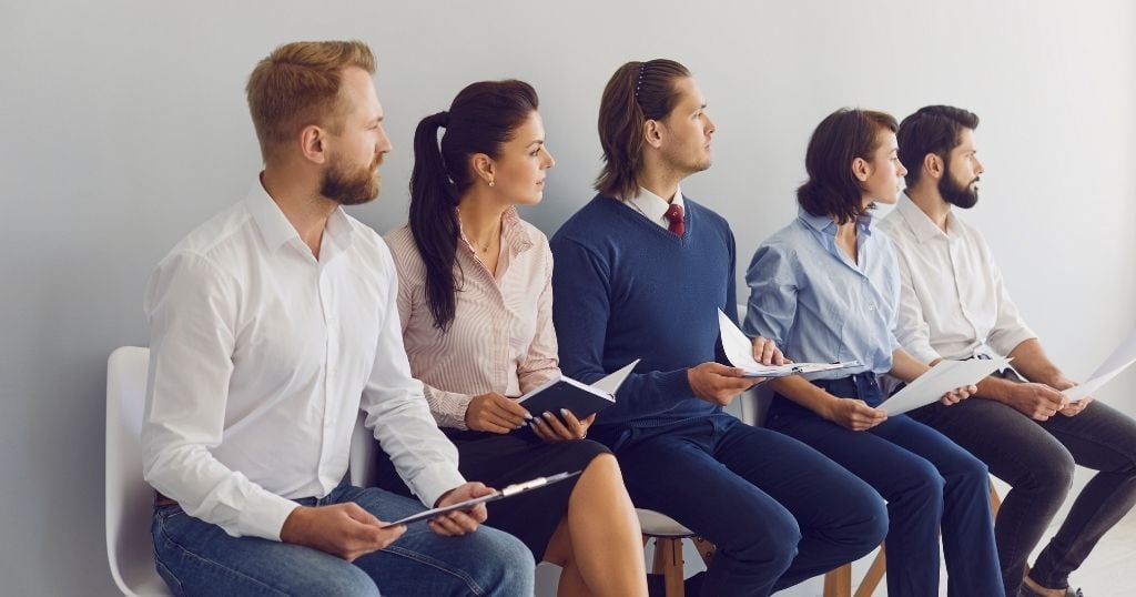 A group of candidates sitting in a row on chairs, holding resumes and notebooks while waiting for an interview.