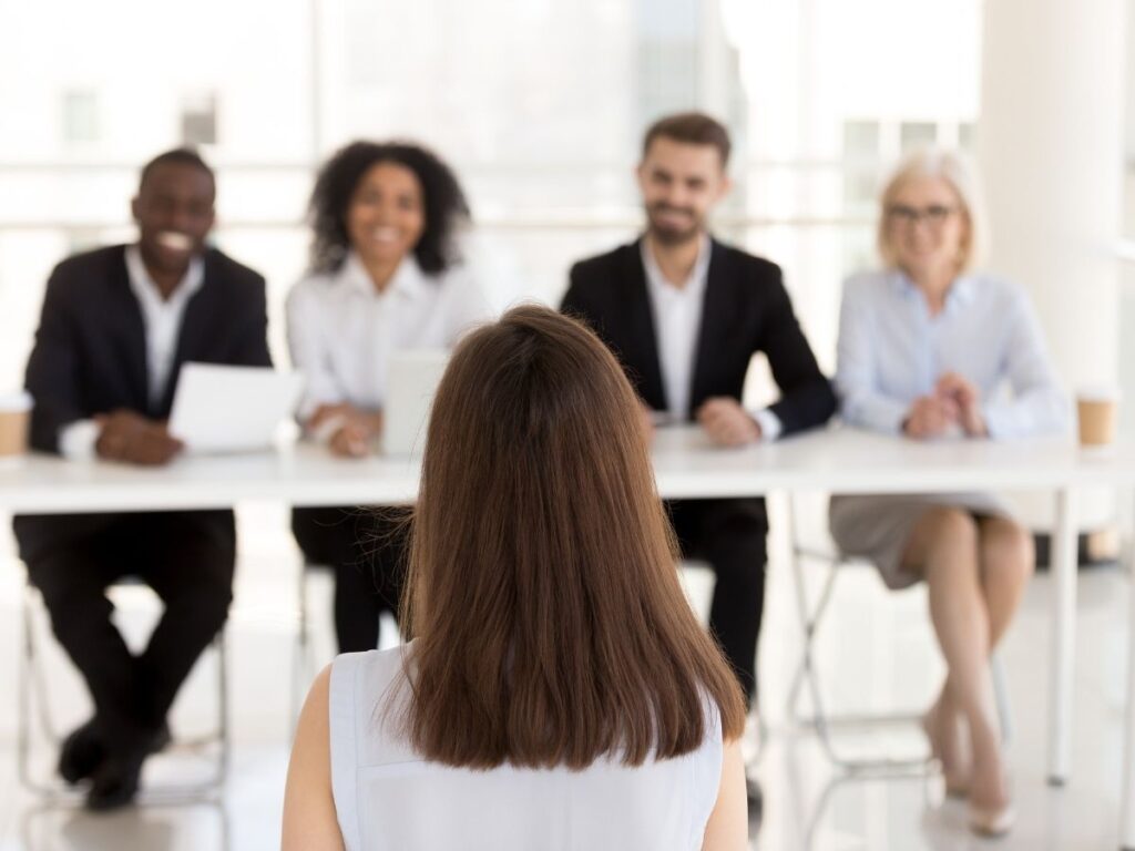 Rear view of a woman facing a panel of four smiling professionals seated at a long white table in a bright office.