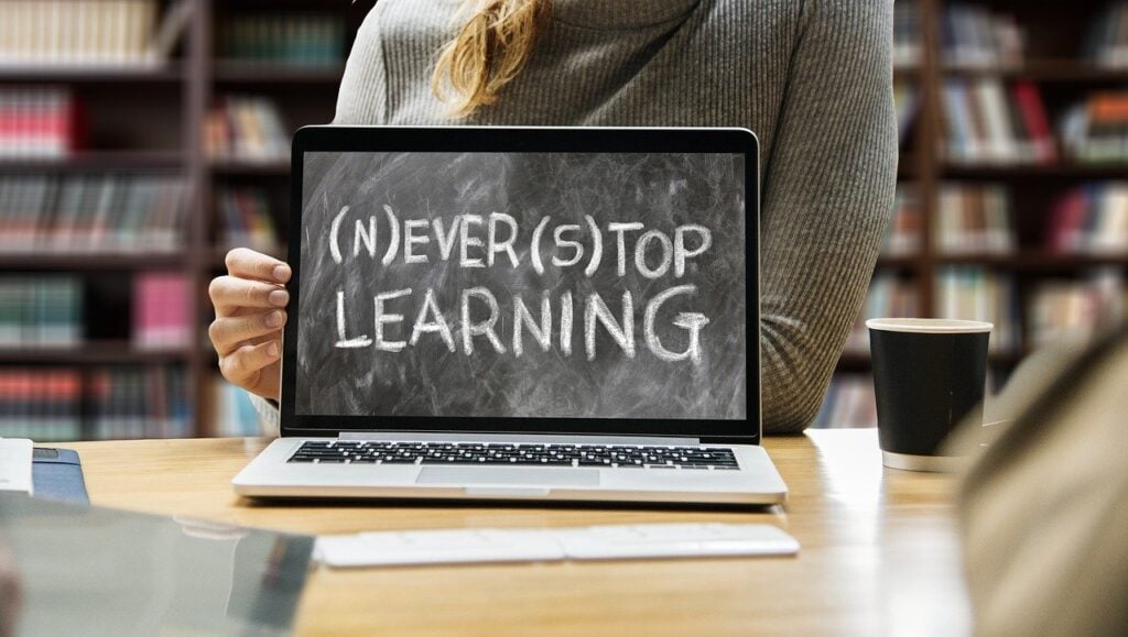 A person holds a laptop displaying a chalkboard graphic with the message "(N)EVER (S)TOP LEARNING"