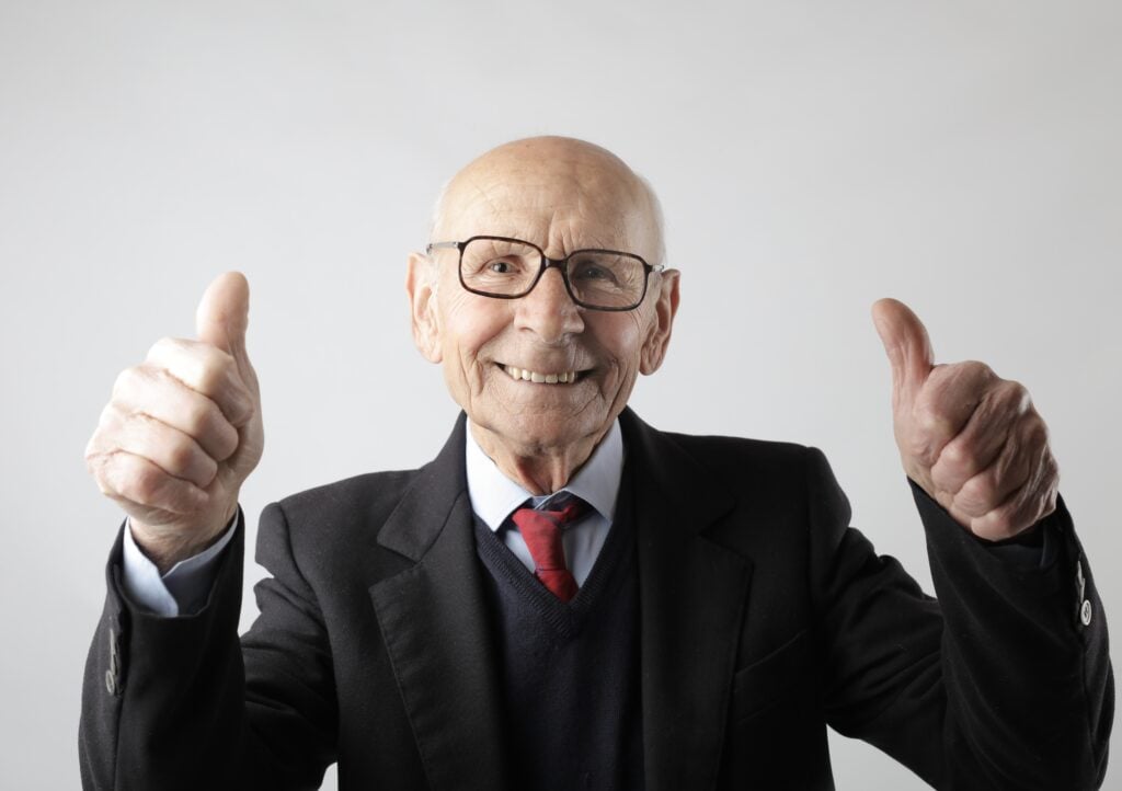 An elderly man in a suit and tie smiling and giving two thumbs up against a plain white background.