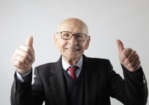 An elderly man in a suit and tie smiling and giving two thumbs up against a plain white background.