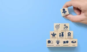 Hand placing a wooden block with 'Leadership' atop a pyramid of blocks with business icons