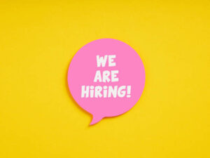 A pink speech bubble on a bright yellow background with the text "WE ARE HIRING!" in white capital letters.