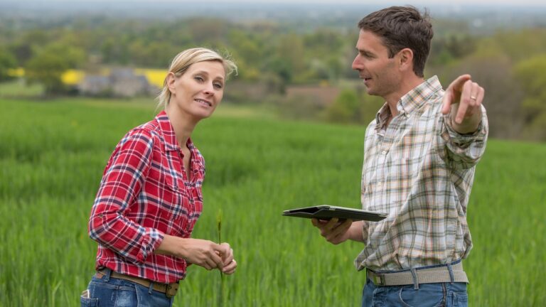 Two farmers, a woman in a red plaid shirt and a man in a tan plaid shirt holding a tablet, discussing crops in a green field.
