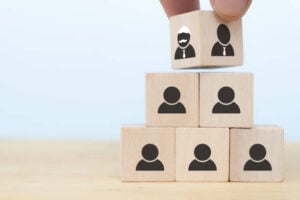 A hand placing a wooden block featuring two leader icons atop a pyramid of blocks with person icons