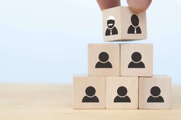 A hand placing a wooden block featuring two leader icons atop a pyramid of blocks with person icons