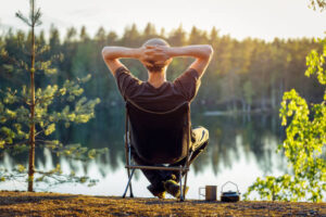 A person sits in a portable camping chair from behind, hands behind their head, looking out over a calm lake at sunset.