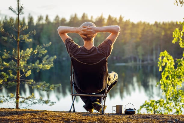 A person sits in a portable camping chair from behind, hands behind their head, looking out over a calm lake at sunset.
