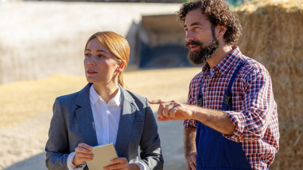 A woman in a professional grey business suit holding a tablet listens to a bearded man as he points toward an outdoor agricultural or farming area.