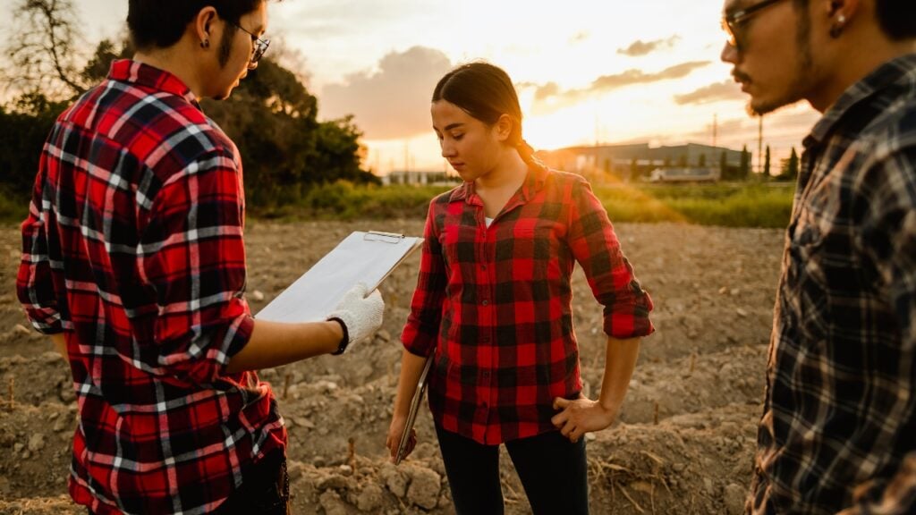 Three people in plaid shirts discussing plans on a farm field at sunset, with one holding a clipboard.