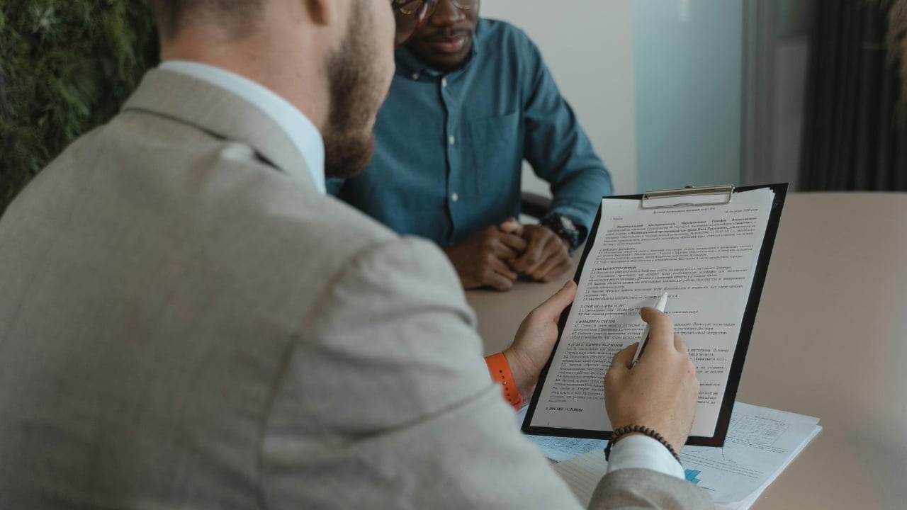 A person in a suit holding a clipboard with a document, pointing to text during a meeting with a colleague.