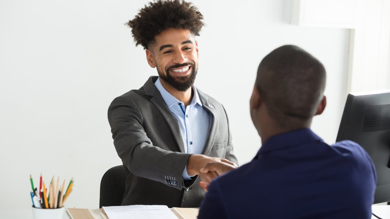 A smiling professional in a suit shaking hands with a client across a desk.